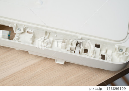 Open white printer and scanner casing interior plastic compartments, spring mechanism and ribbon cable on a wooden desk. Electronics repair, device maintenance, teardown and technical illustration. Open white printer and scanner casing interior plastic compartments, spring mechanism and ribbon cable on a wooden desk. Electronics repair, device maintenance, teardown and technical illustration. 138896199