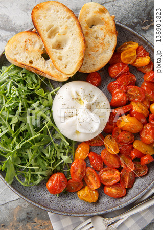 fresh burrata cheese served with roasted cherry tomatoes, green arugula on a dark plate and toasted baguette slices closeup. Vertical top view fresh burrata cheese served with roasted cherry tomatoes, green arugula on a dark plate and toasted baguette slices closeup. Vertical top view 138901823