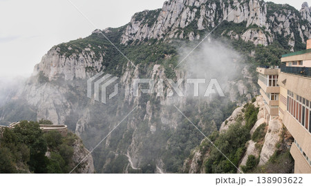 A scenic view of the rugged Montserrat mountains with mist swirling around the peaks and a building perched on the cliffside 138903622