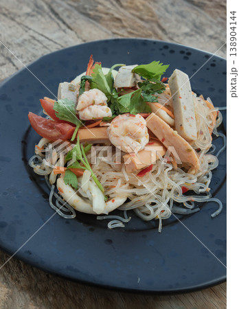 Presented on a matte black plate, this vibrant Yum Woon Sen features shimmering glass noodles tossed with succulent shrimp, tender squid, and savory pork sausage. Aromatic celery and bright tomatoes 138904144