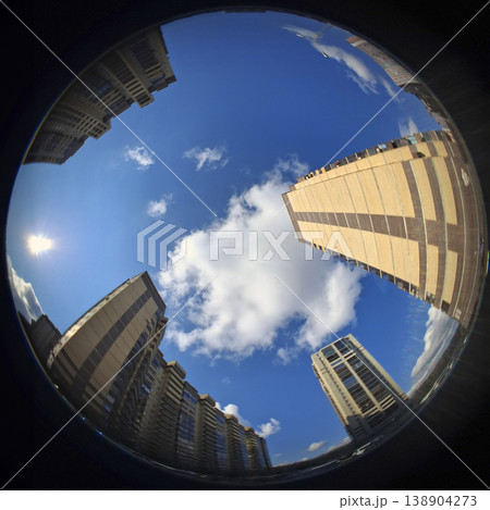 High rise apartment buildings surround a circular view of the sky. The sun shines brightly in the background. Fluffy clouds drift in the blue sky above the urban area in the daytime. High rise apartment buildings surround a circular view of the sky. The sun shines brightly in the background. Fluffy clouds drift in the blue sky above the urban area in the daytime. 138904273
