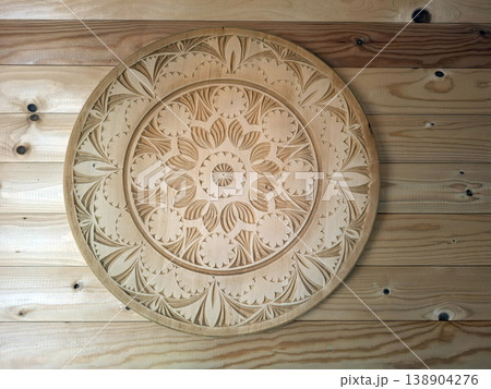 A circular wooden platter with hand carved floral and geometric patterns sits on a wooden wall. This fine craftsmanship shows traditional techniques used in crafting decor items. A circular wooden platter with hand carved floral and geometric patterns sits on a wooden wall. This fine craftsmanship shows traditional techniques used in crafting decor items. 138904276