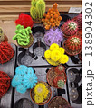 Small cacti in bright colors of blue, yellow, red, and purple are placed in nursery pots. They are arranged neatly on a display shelf in a plant shop. 138904302