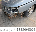 A silver car shows damage on the front corner. The headlight is smashed, and the fender is bent. Rust is evident on the exposed frame. The car appears to be parked on a paved surface. 138904304