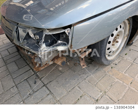 A silver car shows damage on the front corner. The headlight is smashed, and the fender is bent. Rust is evident on the exposed frame. The car appears to be parked on a paved surface. 138904304