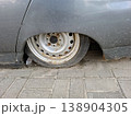 A close up shows a flat tire resting on a rusted steel rim. The vehicle body is low to the ground on a paved surface, highlighting the issue with the wheel. 138904305