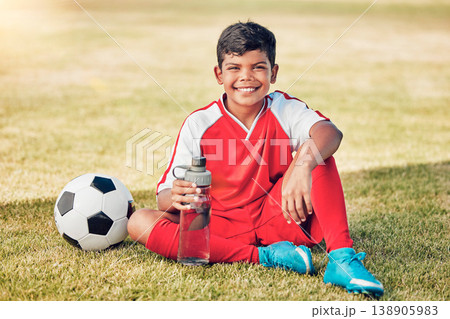 Soccer, children and water with a ball and boy child sitting on a grass pitch or field after training. Football, fitness and hydration with a young male kid at a sports venue, stadium or arena 138905983