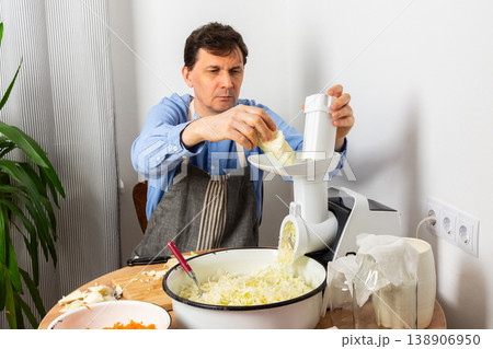 A 53-year-old man prepares cabbage and carrots for fermentation in his kitchen. He uses a machine to shred the vegetables while surrounded by kitchen tools and ingredients A 53-year-old man prepares cabbage and carrots for fermentation in his kitchen. He uses a machine to shred the vegetables while surrounded by kitchen tools and ingredients 138906950