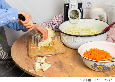 A 53-year-old man cuts cabbage with a knife while preparing to ferment cabbage and carrots. He stands in his kitchen near a vegetable grinder and a bowl of grated carrots A 53-year-old man cuts cabbage with a knife while preparing to ferment cabbage and carrots. He stands in his kitchen near a vegetable grinder and a bowl of grated carrots 138906957