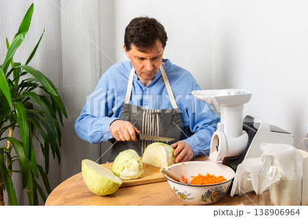 A 53-year-old man cuts cabbage with a knife while preparing to ferment cabbage and carrots. He stands in his kitchen near a vegetable grinder and a bowl of grated carrots A 53-year-old man cuts cabbage with a knife while preparing to ferment cabbage and carrots. He stands in his kitchen near a vegetable grinder and a bowl of grated carrots 138906964