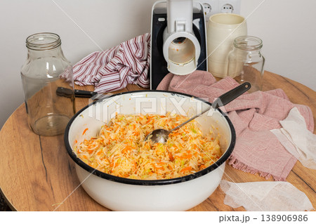 Cabbage and carrots are mixed together for fermentation in a bowl on a wooden table. Jars and kitchen equipment are present in the background, showing a food preparation scene Cabbage and carrots are mixed together for fermentation in a bowl on a wooden table. Jars and kitchen equipment are present in the background, showing a food preparation scene 138906986