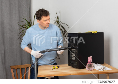 A 53-year-old man cleans dust from a computer system unit with a vacuum cleaner in a tidy room. He carefully removes dirt to keep the device running well A 53-year-old man cleans dust from a computer system unit with a vacuum cleaner in a tidy room. He carefully removes dirt to keep the device running well 138907027