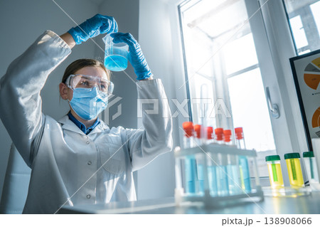 A researcher wearing a lab coat, gloves, mask, and goggles analyzes a blue solution in a beaker. The laboratory setting highlights scientific research, innovation, and chemical analysis A researcher wearing a lab coat, gloves, mask, and goggles analyzes a blue solution in a beaker. The laboratory setting highlights scientific research, innovation, and chemical analysis 138908066