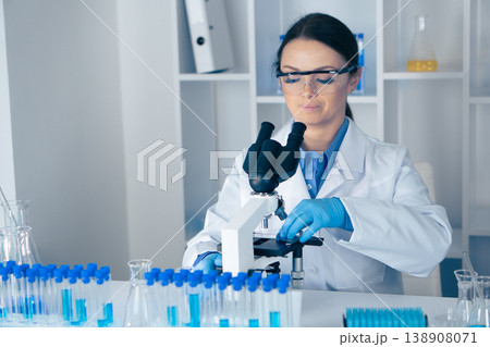 A dedicated female scientist examines samples through a microscope, surrounded by test tubes and advanced lab equipment, utilizing the latest technology for research and discovery A dedicated female scientist examines samples through a microscope, surrounded by test tubes and advanced lab equipment, utilizing the latest technology for research and discovery 138908071
