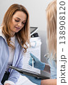 Female patient attentively listens to a doctor explaining medical information using a tablet in a bright clinic. Healthcare provider wears gloves, emphasizing safety and professionalism 138908120