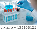 A close up view of laboratory research shows a gloved hand handling a pipette with blue liquid. Test tubes, beakers, and scientific equipment are visible in a clean, bright lab setting 138908122