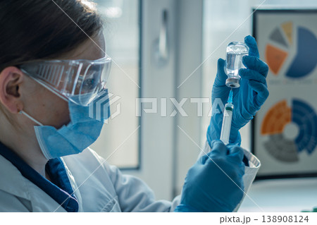 Doctor or nurse wearing blue gloves and lab coat fills syringe from glass vial, focusing on vaccination process. Modern healthcare and immunization concept in bright hospital environment 138908124