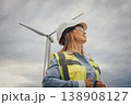 A highly skilled female engineer, dressed in safety gear, stands confidently before a towering wind turbine, which symbolizes both progress and innovation in the vital field of renewable energy 138908127