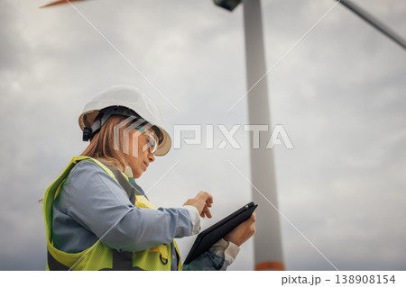 A qualified female engineer in safety gear inspects a wind turbine with a tablet, showcasing the link between technology and sustainable renewable energy practices vital for our future A qualified female engineer in safety gear inspects a wind turbine with a tablet, showcasing the link between technology and sustainable renewable energy practices vital for our future 138908154