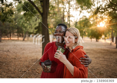 Two happy multi-ethnic friends laughing and hugging in a park at sunset, drinking coffee 138908369