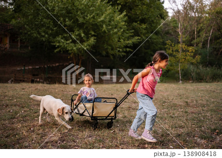 Girl pulling her twin sister in a cart with their dog running alongside Girl pulling her twin sister in a cart with their dog running alongside 138908418