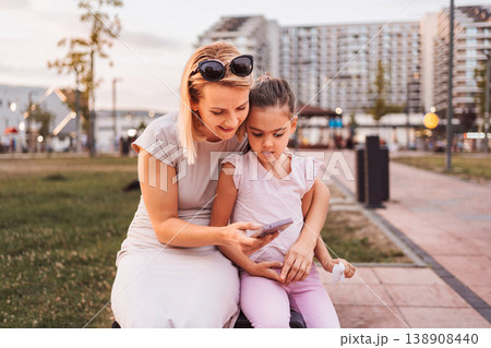 Mother and daughter looking at smartphone sitting on bench in urban park 138908440