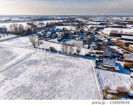High-angle aerial view of a snowy European village and industrial zone. Residential houses and fields covered in fresh white snow under a bright winter sun. Scenic cold weather landscape. High-angle aerial view of a snowy European village and industrial zone. Residential houses and fields covered in fresh white snow under a bright winter sun. Scenic cold weather landscape. 138909048