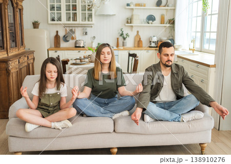 Yoga mindfulness meditation emotional wellbeing. Happy family practicing yoga at home Mother father daughter sitting in lotus pose meditating relaxing on couch Family doing breathing practice together Yoga mindfulness meditation emotional wellbeing. Happy family practicing yoga at home Mother father daughter sitting in lotus pose meditating relaxing on couch Family doing breathing practice together 138910276