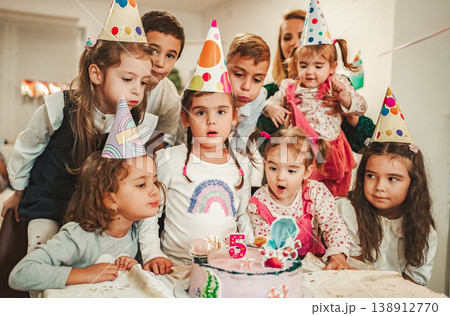 Group of children gather to celebrate a birthday with hats and a cake while preparing to blow out candles Group of children gather to celebrate a birthday with hats and a cake while preparing to blow out candles 138912770