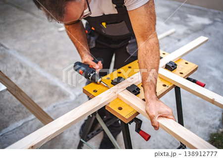 Mature male carpenter using electric screwdriver on a wooden plank outdoors Mature male carpenter using electric screwdriver on a wooden plank outdoors 138912777