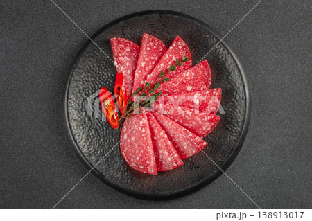 Sliced salami arranged on black plate with chili and herbs top view 138913017