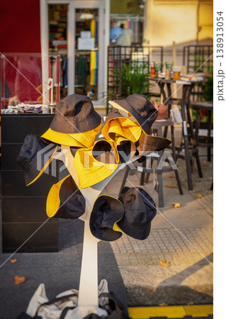 Display of bucket hats in black and yellow on stand outside street shop with clothing store background and outdoor retail setting. Fashion retail, street market, summer accessory, urban shopping Display of bucket hats in black and yellow on stand outside street shop with clothing store background and outdoor retail setting. Fashion retail, street market, summer accessory, urban shopping 138913054
