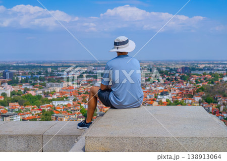 Young man sitting on concrete ledge overlooking city skyline with buildings, roofs and streets under sky, travel scene with urban perspective. Concept of travel, tourism lifestyle, reflections on life Young man sitting on concrete ledge overlooking city skyline with buildings, roofs and streets under sky, travel scene with urban perspective. Concept of travel, tourism lifestyle, reflections on life 138913064