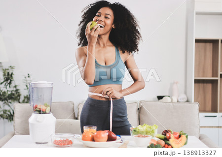 Young nutritionist showing bell peppers during an online video call consulting about healthy eating, sitting at home at a table with vegetables, laptop and measuring tape 138913327