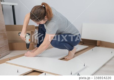 A Caucasian woman uses a screwdriver to tighten a screw while assembling a cabinet. 138914220