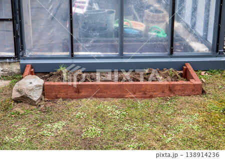 Preparing Wooden Raised Garden Bed Along Greenhouse Wall in Backyard 138914236