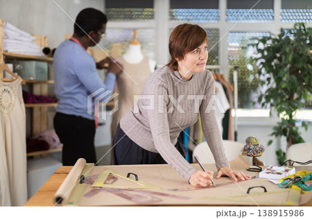 Woman draw dress pattern of product on paper. Companion work in background with mannequin Woman draw dress pattern of product on paper. Companion work in background with mannequin 138915986