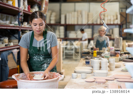 Young female worker in pottery factory lowers fired clay plates and cups into container with glaze 138916014