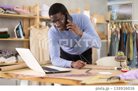Tailor communicating on phone and reviewing specifications on laptop in workshop 138916016