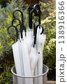 White umbrellas with black handles stored in a metal mesh stand, set against lush green garden shrubs. Clean, minimal, and elegant 138916366