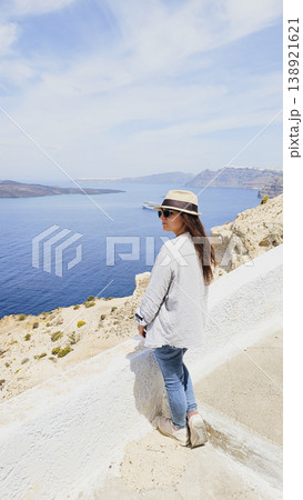 Stylish woman wearing straw hat and linen shirt overlooking Santorini caldera with Aegean Sea panorama and iconic Greek architecture Stylish woman wearing straw hat and linen shirt overlooking Santorini caldera with Aegean Sea panorama and iconic Greek architecture 138921621