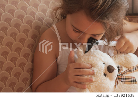 Playing doctor: a small girl examines a teddy bear's ear with an otoscope Playing doctor: a small girl examines a teddy bear's ear with an otoscope 138921639