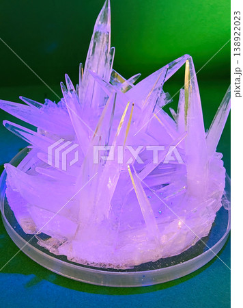 Close up view of sharp, translucent crystals forming in a petri dish under purple and green lighting, showcasing a scientific experiment 138922023