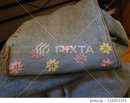 Denim cuff featuring hand-stitched pastel floral embroidery on a rolled hem, showcasing detailed needlework, texture and casual personalized fashion. Ideal for DIY, upcycled clothing, craft projects 138923305