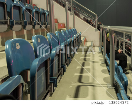 Modern blue stadium seats stand mostly empty with a solitary figure quietly using a phone, representing contemplation and anticipation at a public event 138924572