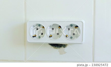 A front-view minimalist shot of a white triple electrical wall socket. Mounted on a clean white tiled wall with visible installation detail. 138925472