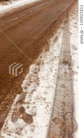 A textured view of a muddy dirt road partially covered with slushy white snow, tire tracks, and footprints. 138925811