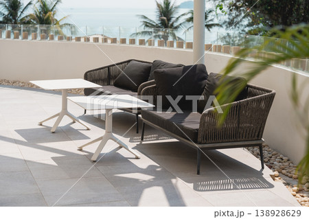 Modern Outdoor Lounge Area with Rattan Sofa and Minimalist Table Next to Tropical Scenery and Ocean View in Bright Daylight Modern Outdoor Lounge Area with Rattan Sofa and Minimalist Table Next to Tropical Scenery and Ocean View in Bright Daylight 138928629