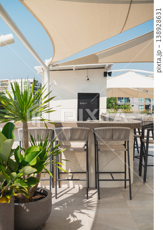 Modern Outdoor Bar with Stylish Seating, Tropical Plants, and Shade Canopy in a Bright Sunny Day at a Contemporary Rooftop Venue Modern Outdoor Bar with Stylish Seating, Tropical Plants, and Shade Canopy in a Bright Sunny Day at a Contemporary Rooftop Venue 138928631