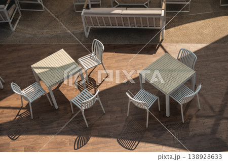Modern Outdoor Dining Area with White Tables and Chairs, Minimalist Design, Warm Sunlight Casting Shadows on Wooden Floor 138928633